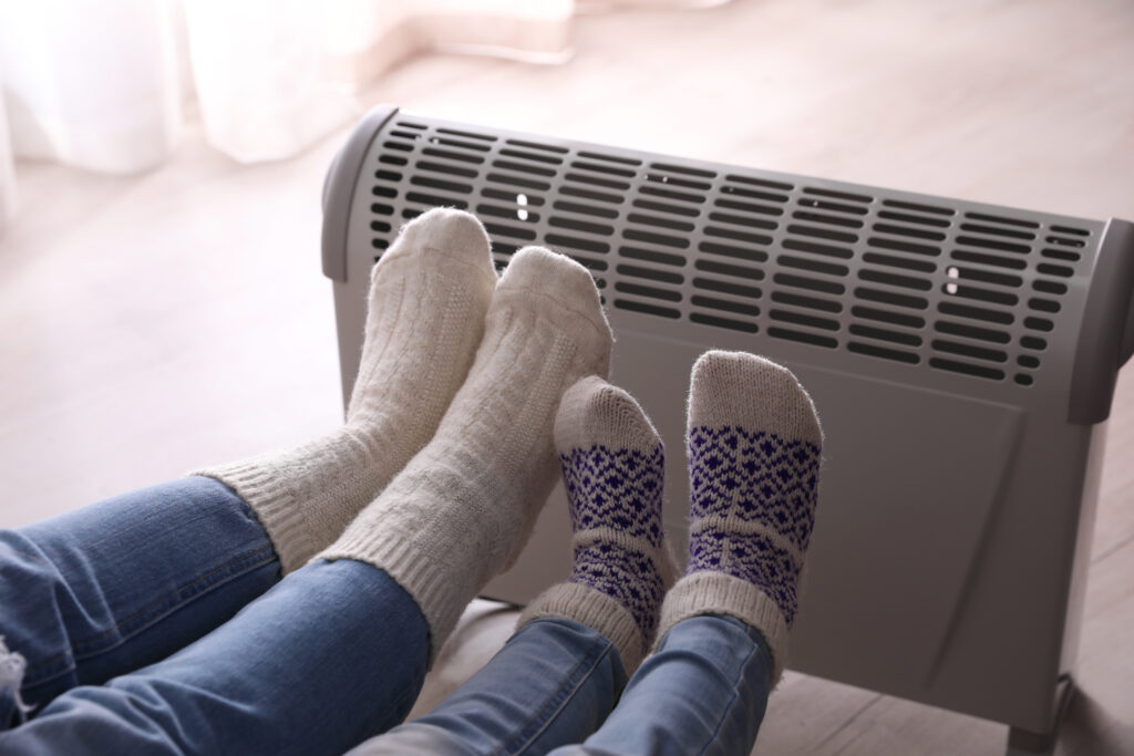 Two people wearing warm socks sit near a heater, illustrating how cold floors and poor insulation can impact home comfort and energy efficiency.